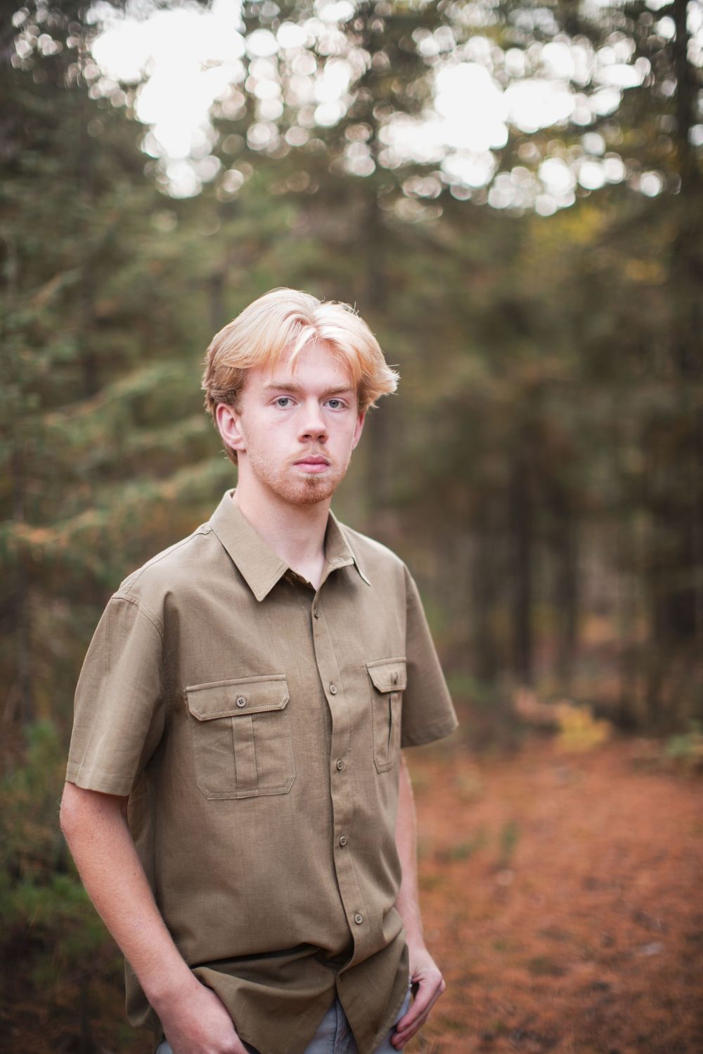 A person in a tan uniform shirt poses in an autumn forest setting with fallen leaves on the ground.