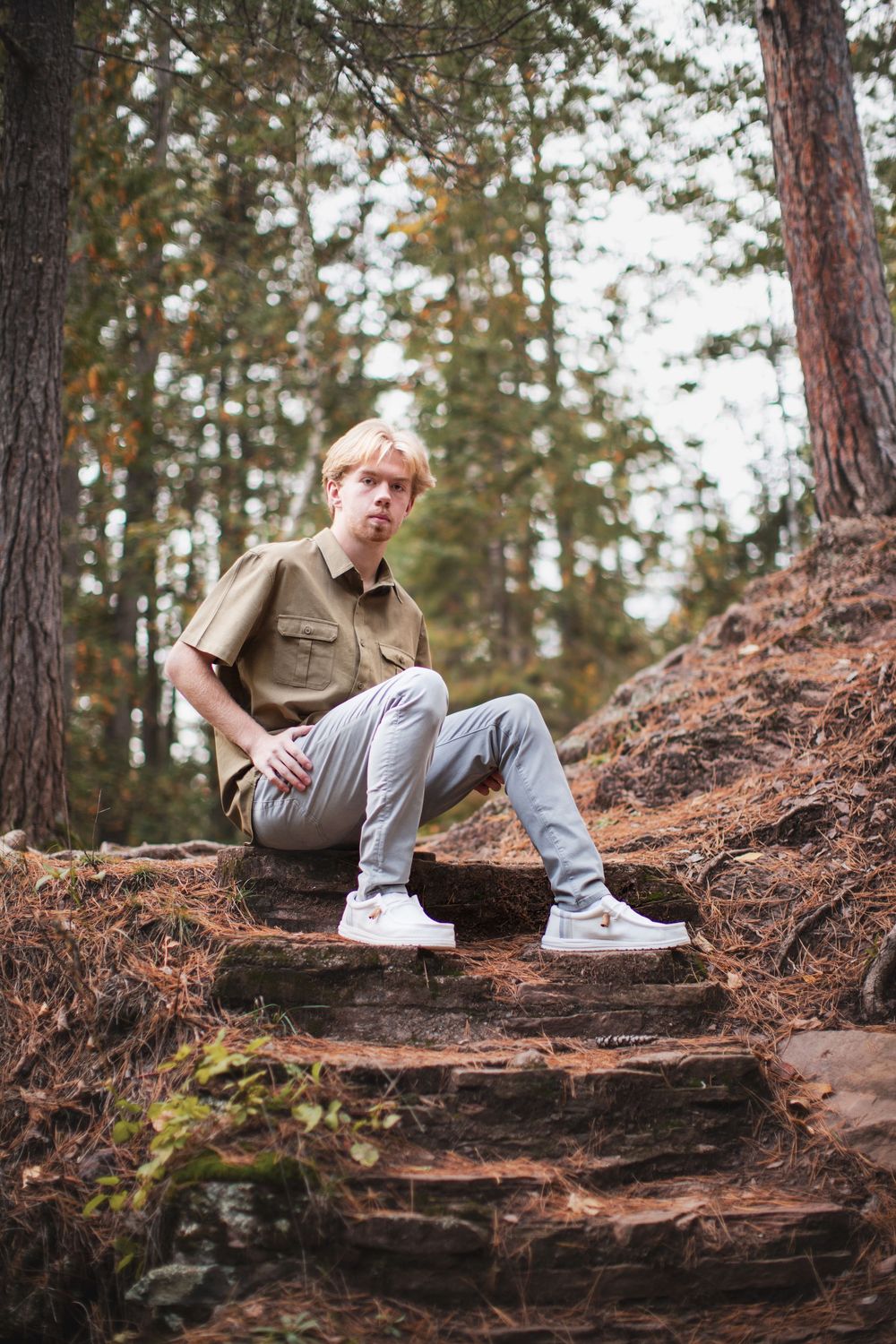 A person in casual attire sits on a fallen log in a forest setting surrounded by pine trees and fallen leaves.