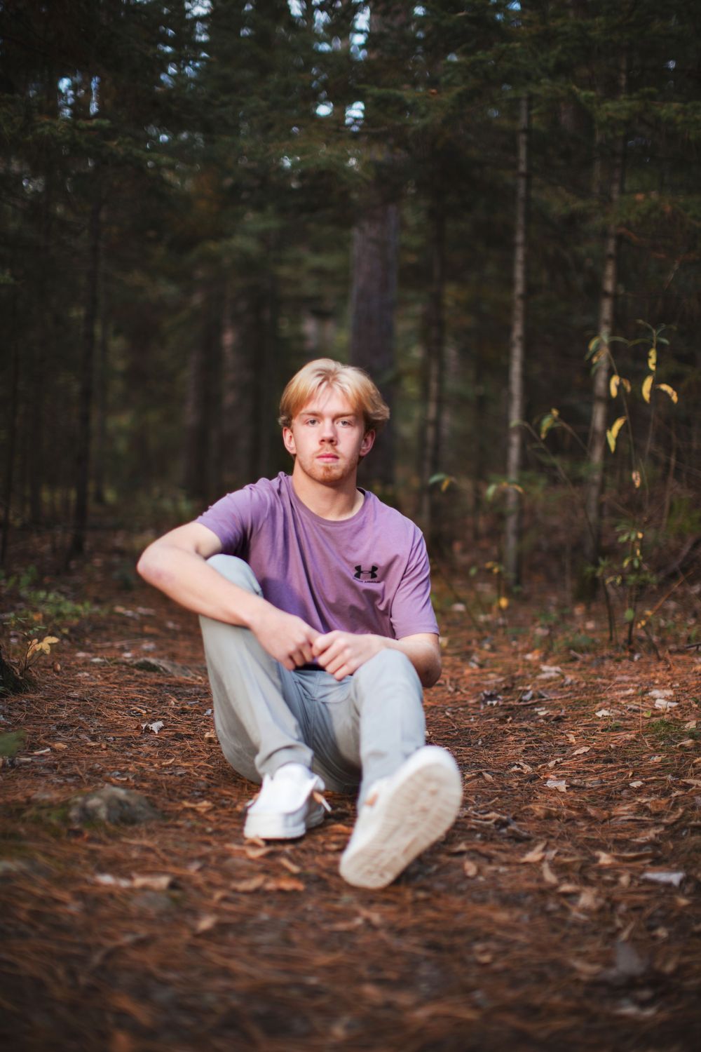 A relaxed outdoor scene showing someone in a purple t-shirt sitting on forest ground covered in pine needles.