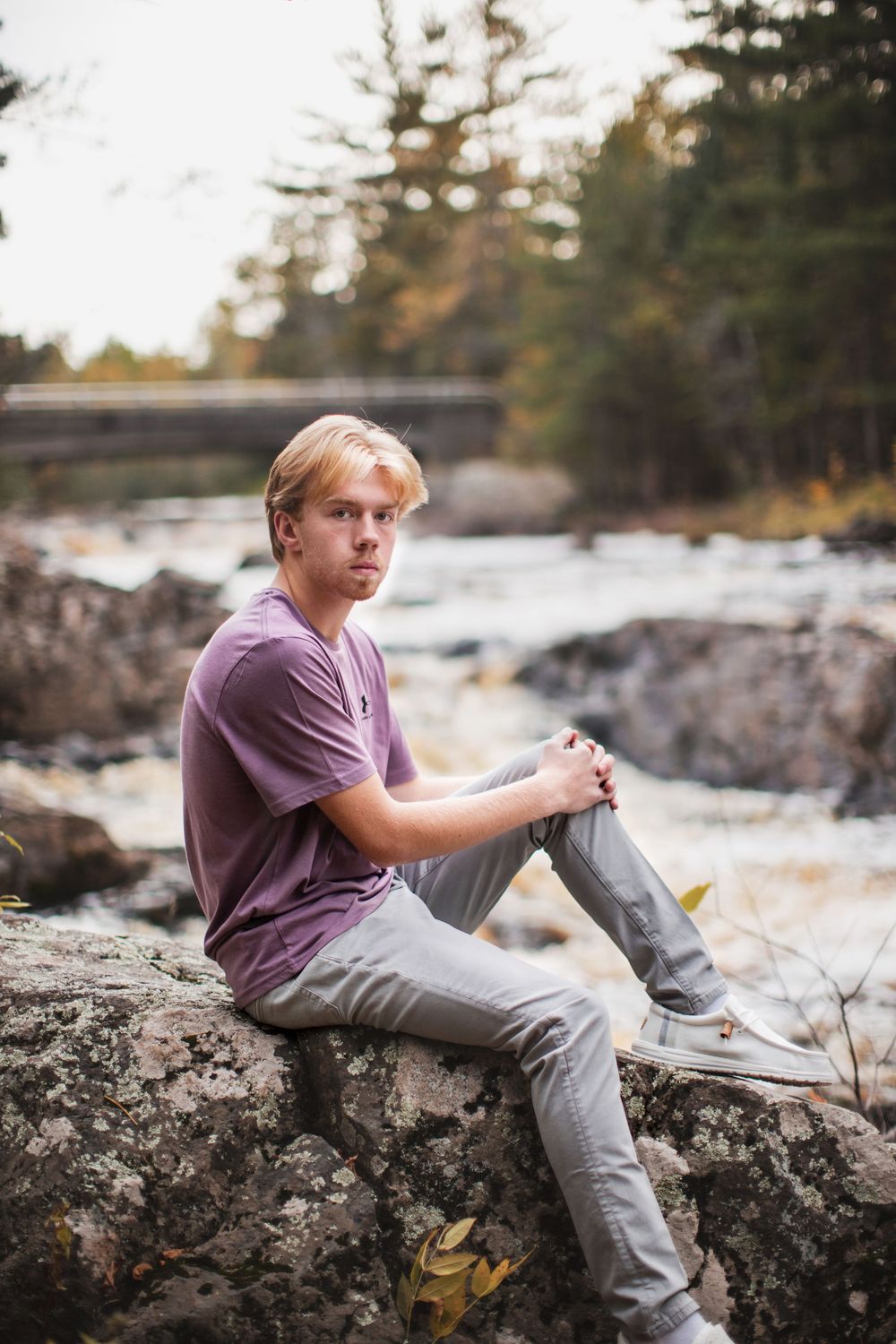 Person in purple shirt sits contemplatively on rocky riverbank during autumn afternoon.
