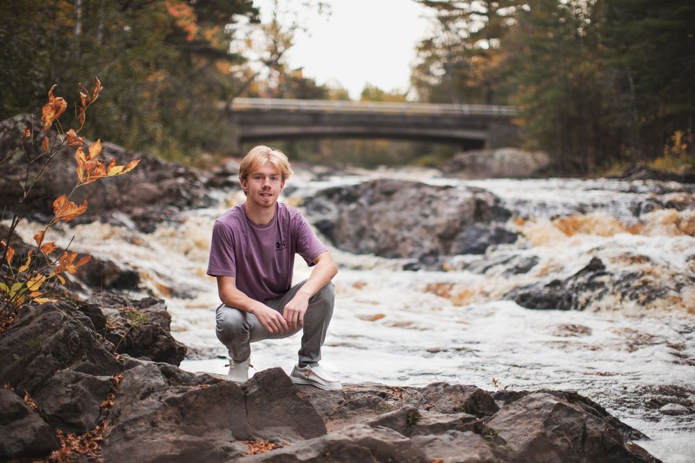 A person in casual clothing poses near a rocky riverbed during autumn with colorful fall foliage in the background.