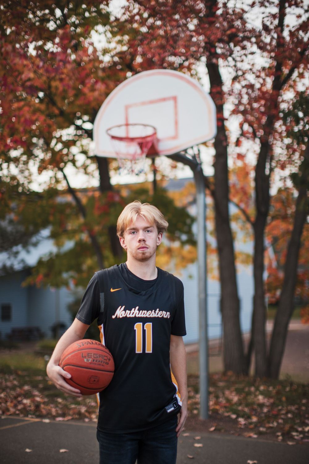 Basketball player in black jersey stands beneath autumn trees with red and orange leaves near outdoor court.