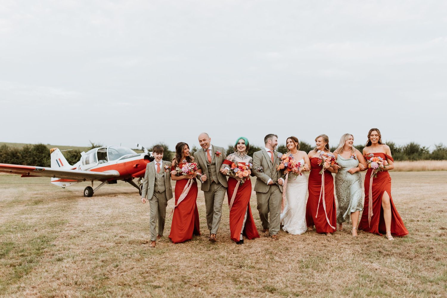 Wedding party in red and beige dresses poses together in a rustic outdoor setting with a small airplane in background.
