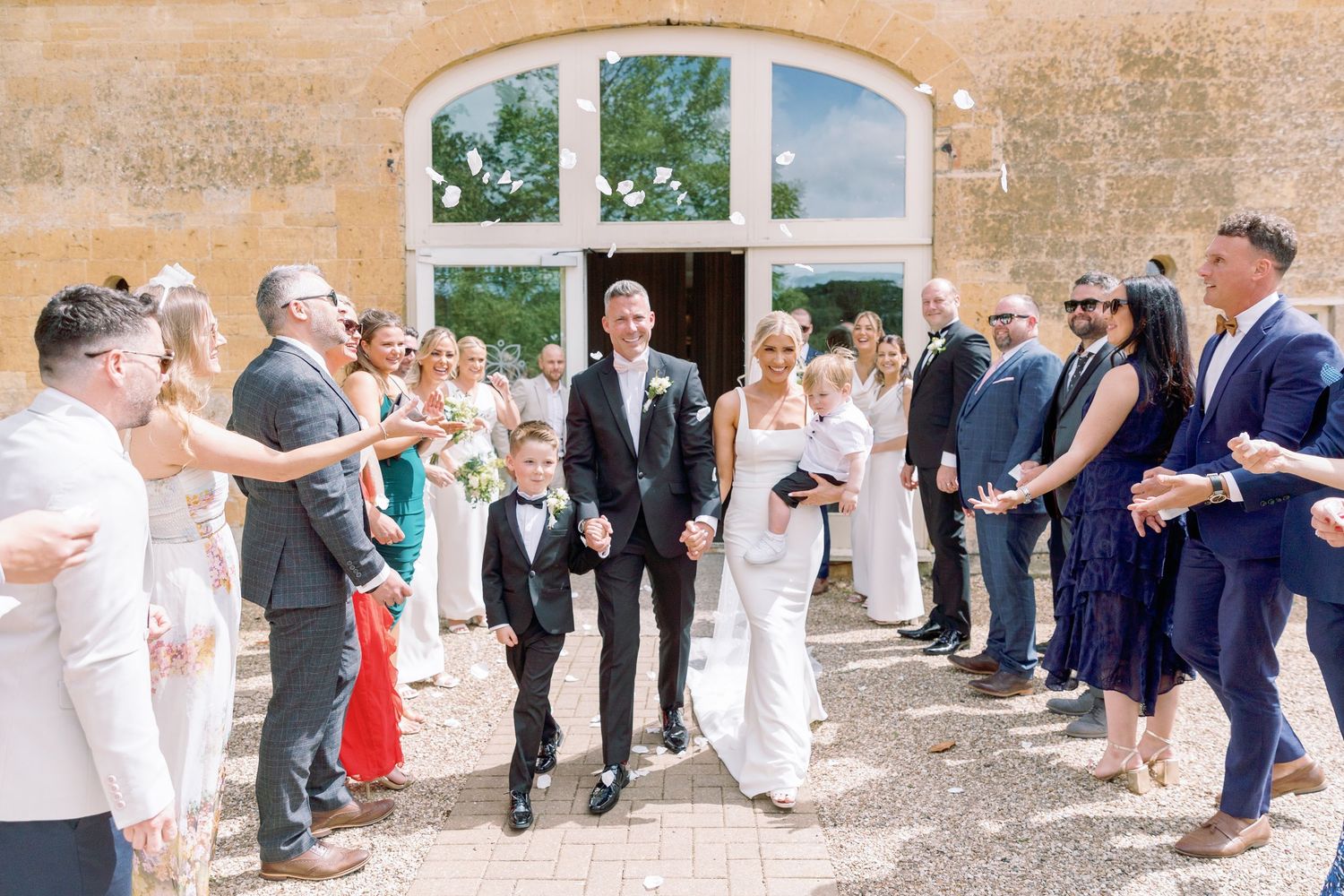 Wedding guests line up along a stone wall to throw confetti at a couple during an outdoor celebration.