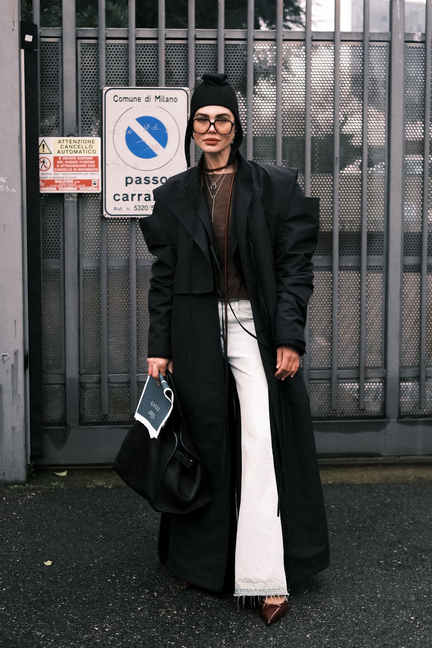 Street style fashion with long black coat, white flare pants, beanie and glasses against industrial gate background.