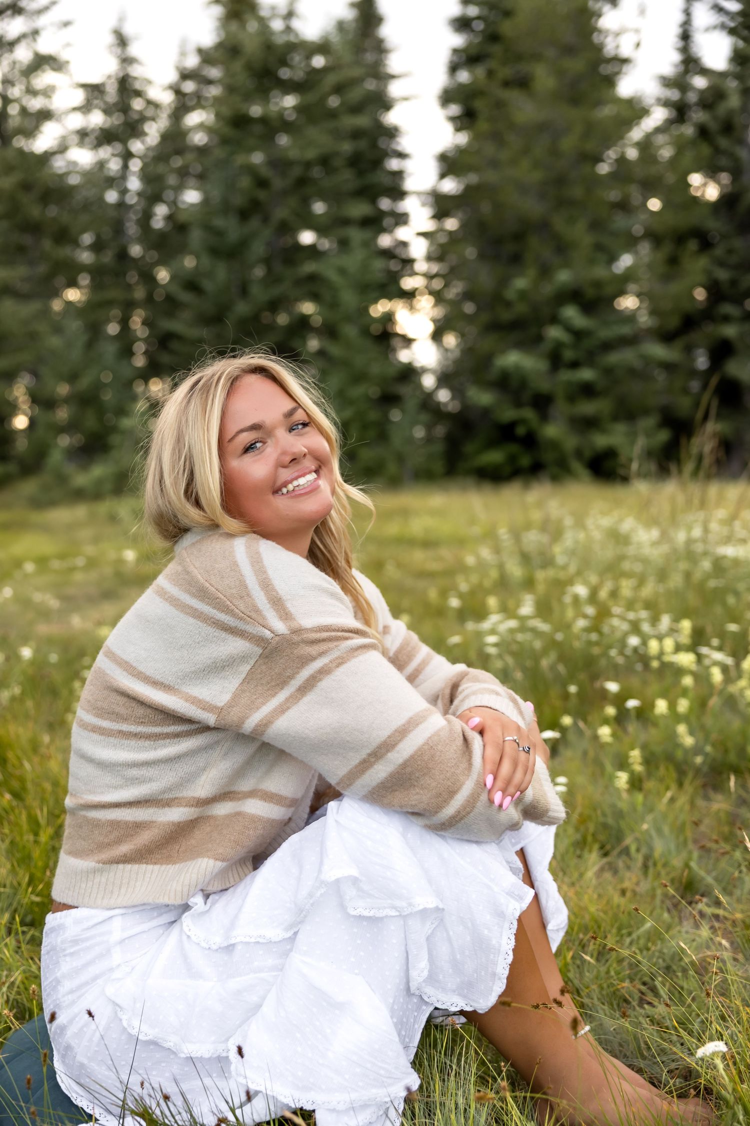 A person in a cream sweater sits in a wildflower meadow at sunset among evergreen trees.