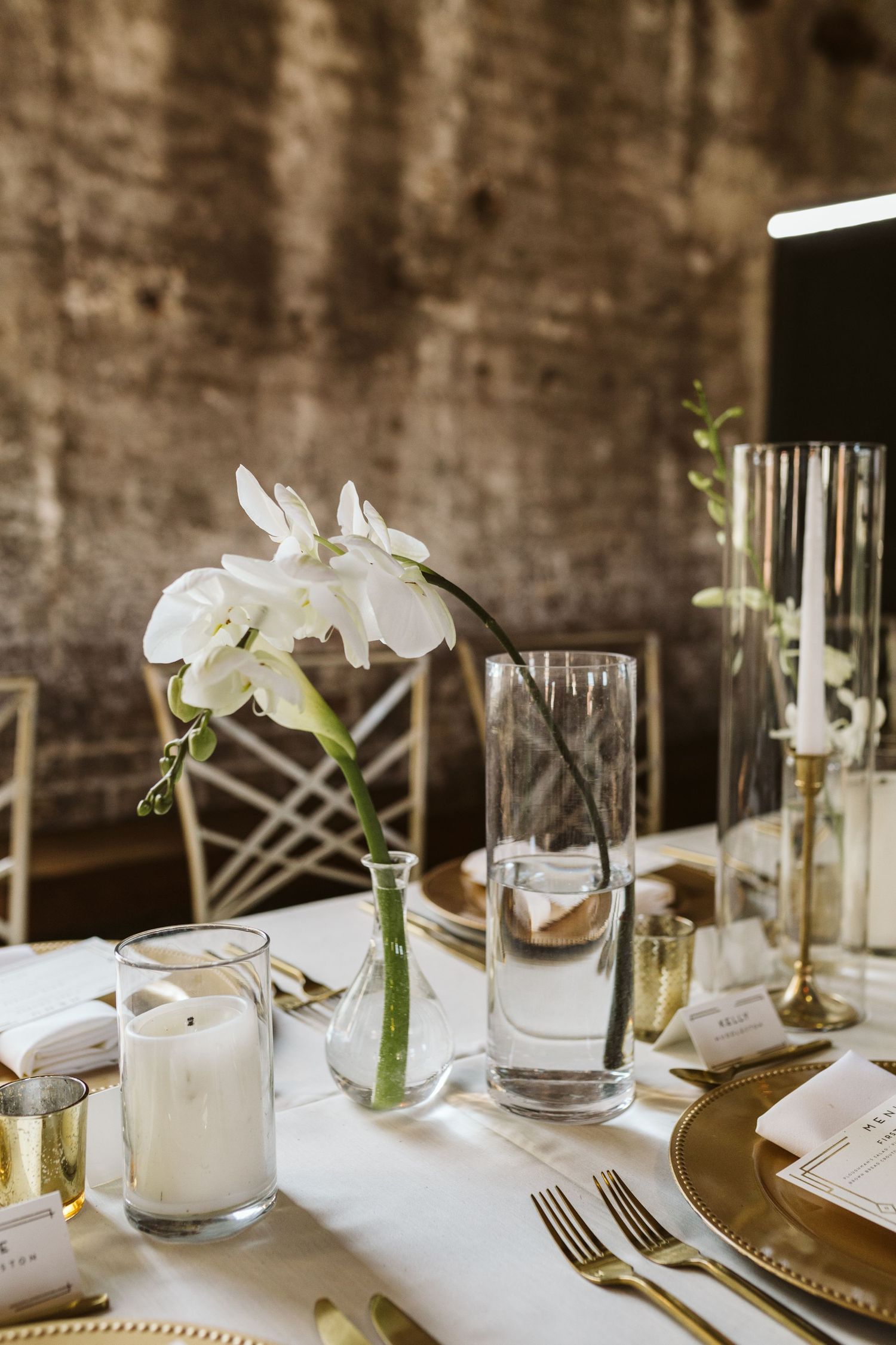 Elegant table setting with white orchids, glass vases, and place settings against a rustic brick wall backdrop.