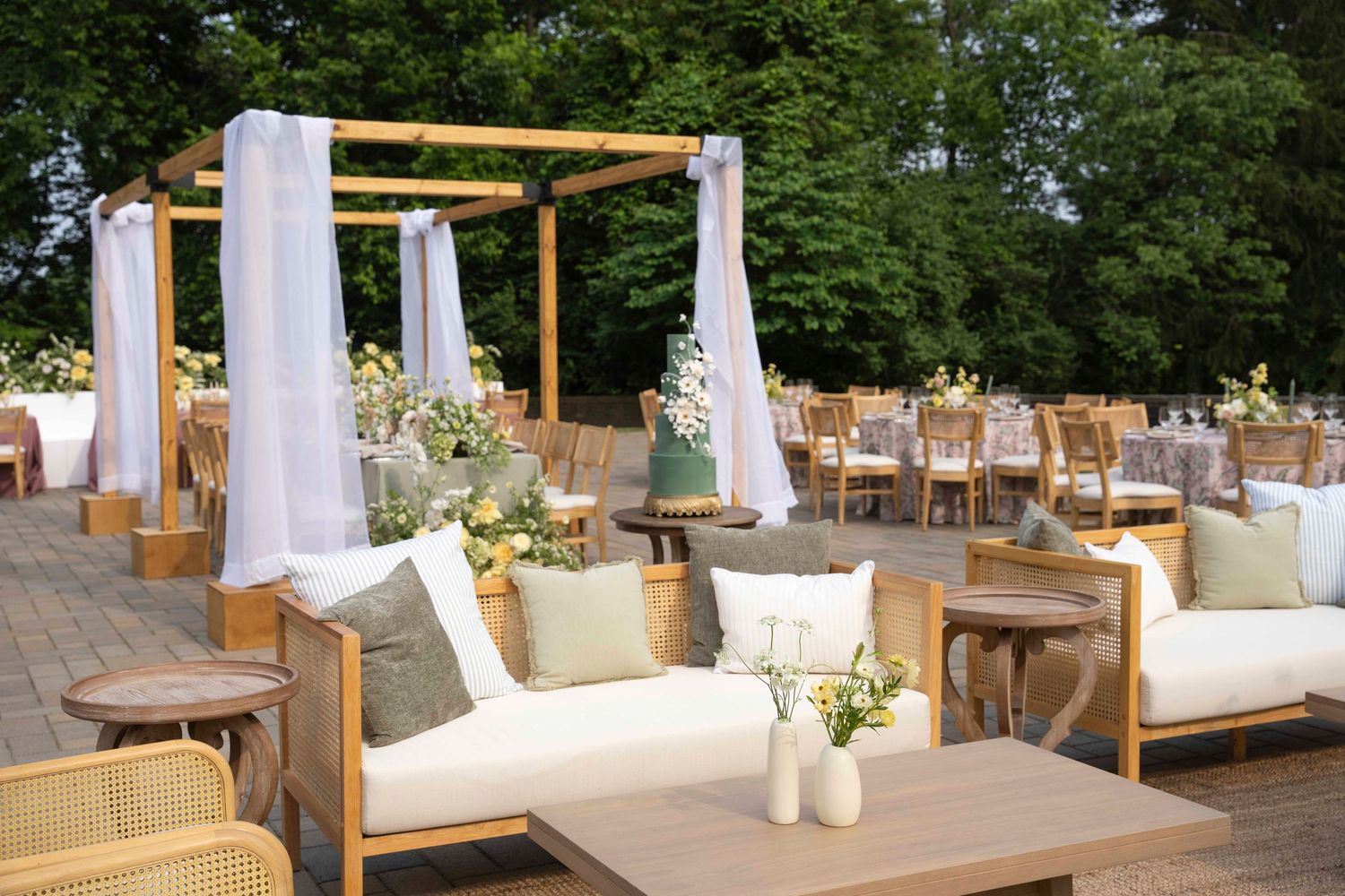 Elegant outdoor lounge area with white fabric drapes, wooden furniture, and natural greenery backdrop for events.