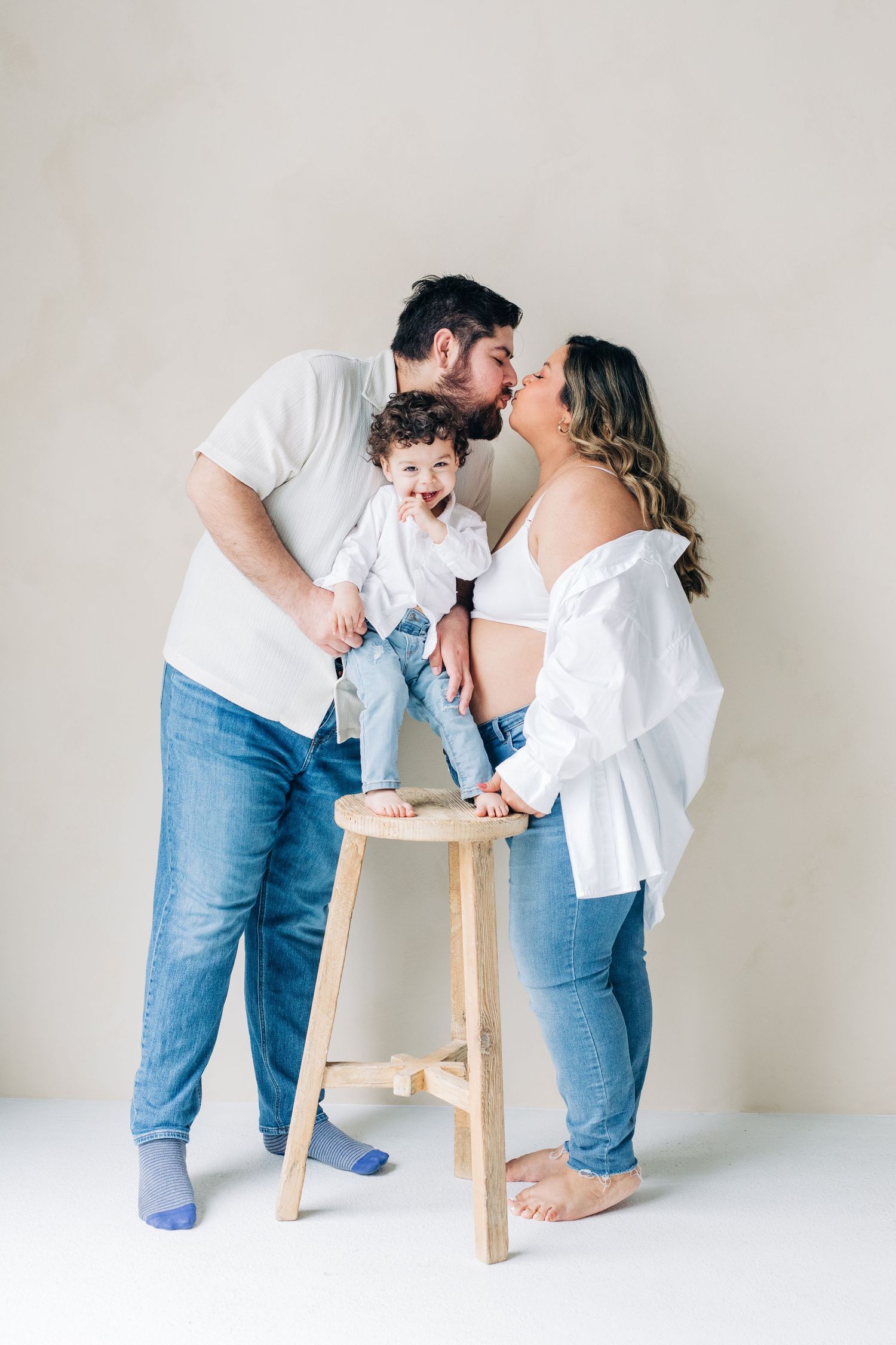 Family in matching white shirts and jeans share sweet moments with their baby on a wooden stool against neutral backdrop.
