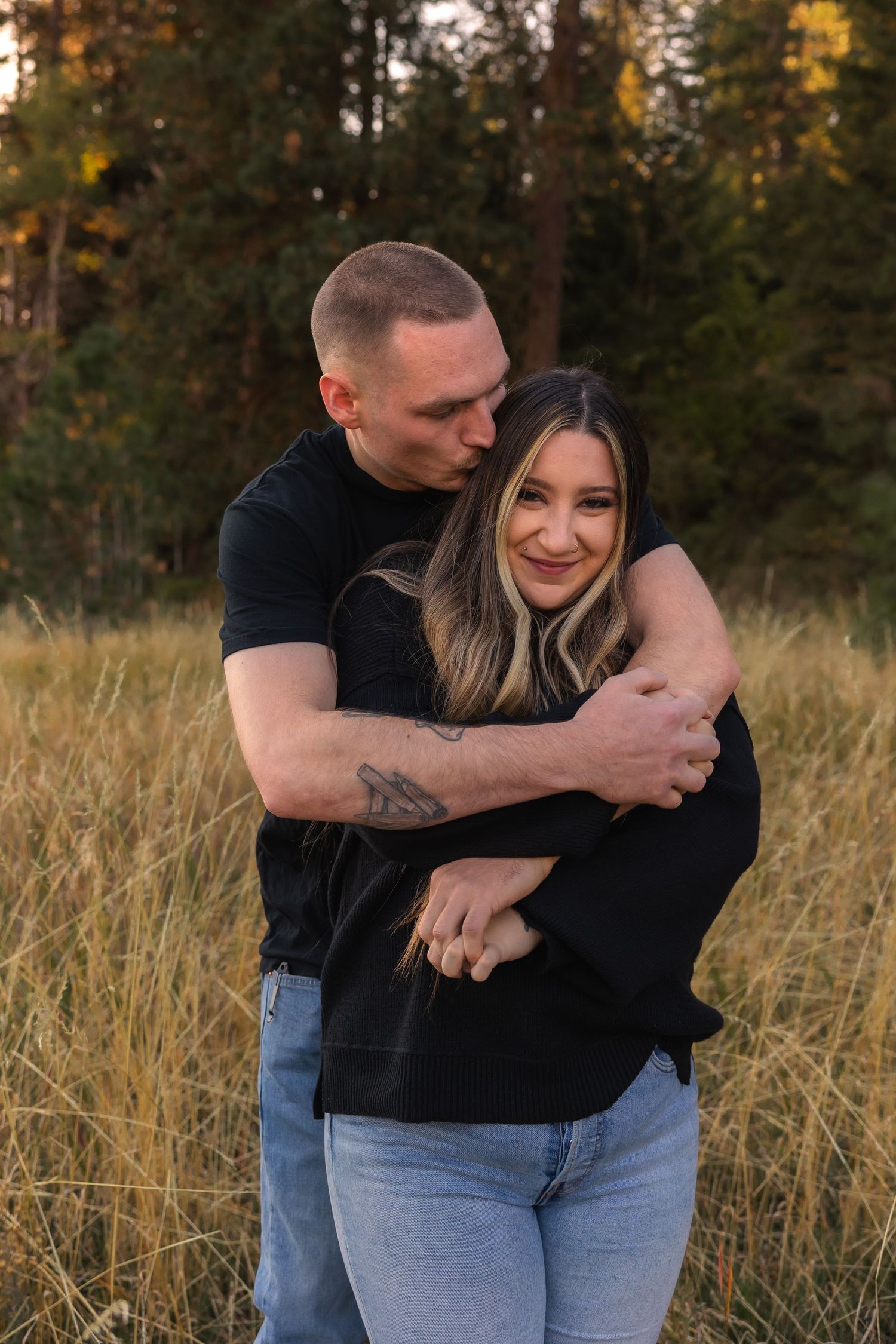 A couple embraces in a field of tall grass during golden hour with autumn trees in the background.