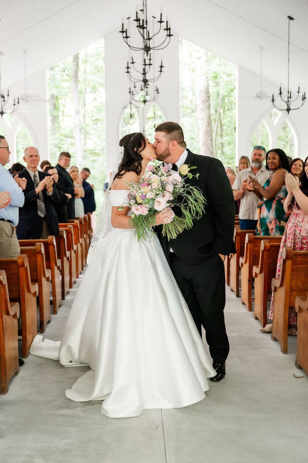 Wedding ceremony in a chapel with chandeliers as guests watch a couple share their first kiss.