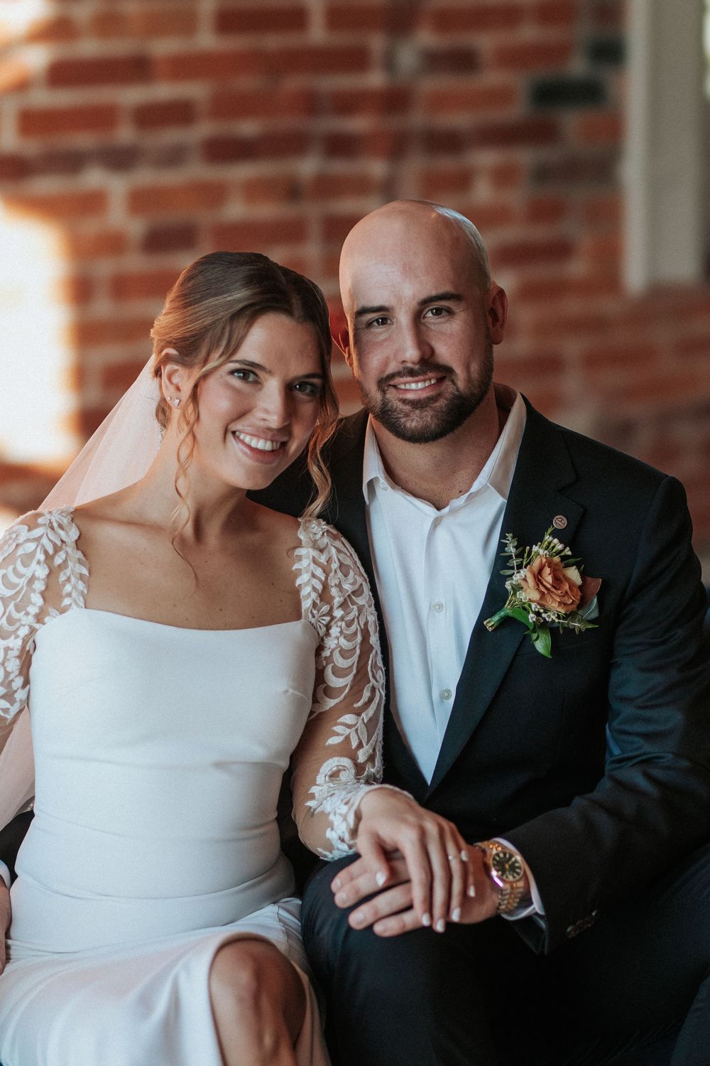 Elegant wedding couple in white dress and black tuxedo sharing an intimate moment against brick wall.