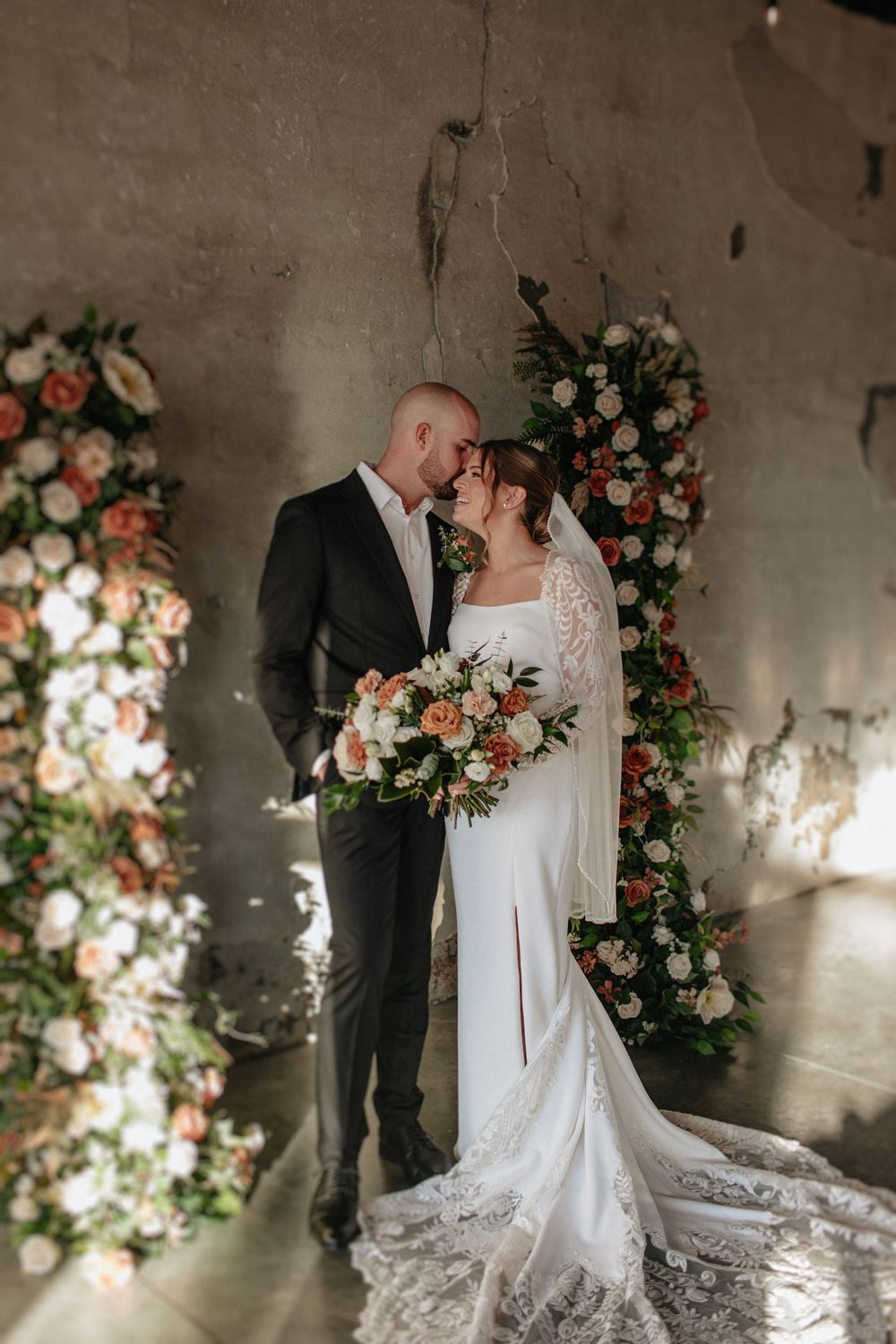 Wedding couple posing by floral arrangements with peach and white blooms against concrete wall.