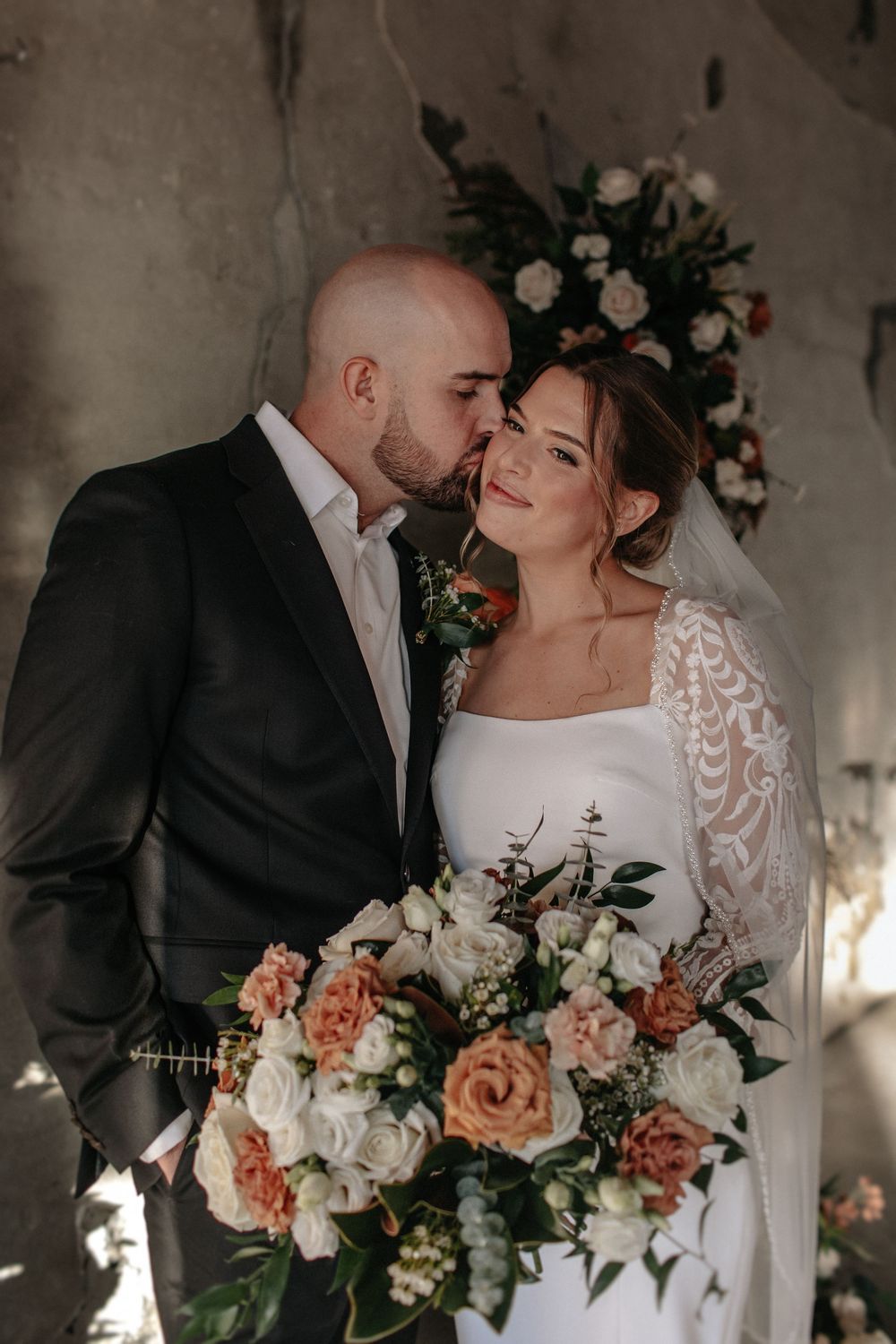 Romantic wedding portrait with peach and white floral arrangements against industrial backdrop.