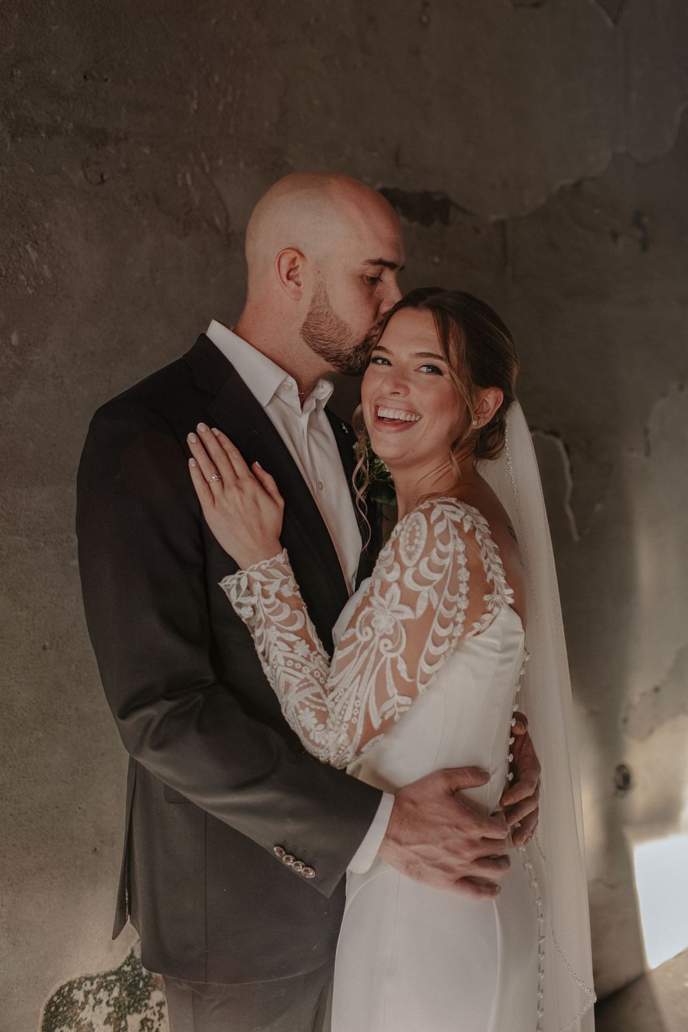 Intimate wedding portrait moment captured against textured concrete wall with natural lighting.