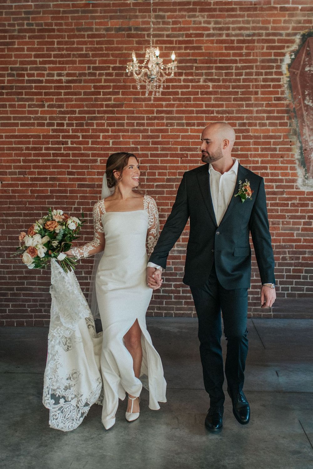 Wedding couple walking together showing details of lace dress and classic tuxedo against brick wall.