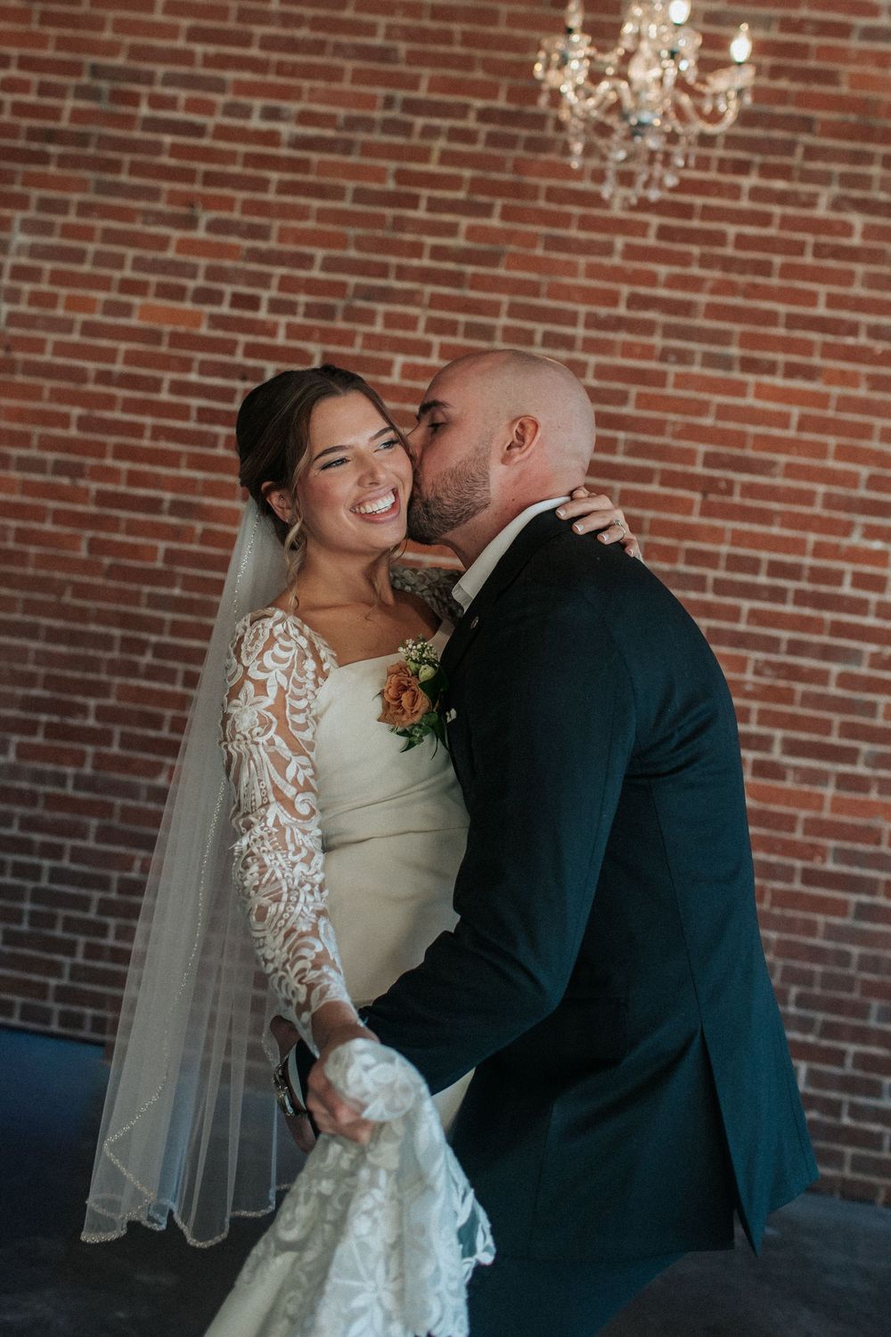 A tender wedding moment captured against a rustic brick wall backdrop with elegant chandeliers overhead.