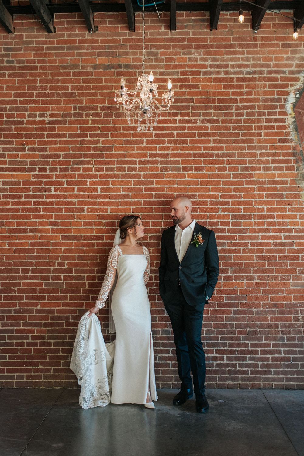 Wedding couple poses together against an industrial brick wall with crystal chandeliers in the background.