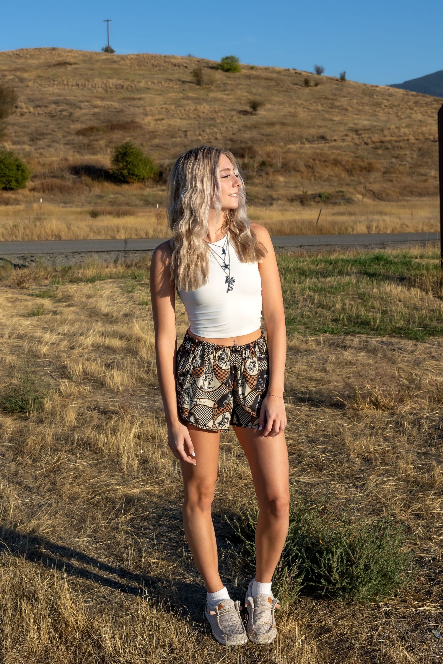 Person wearing patterned shorts and white top poses in a grassy field against dry hillside backdrop.