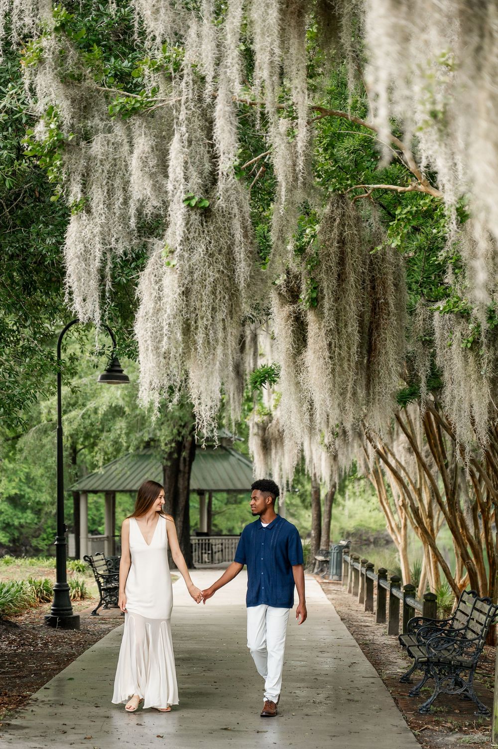 Couple walks hand in hand under Spanish moss-draped trees along a park path.
