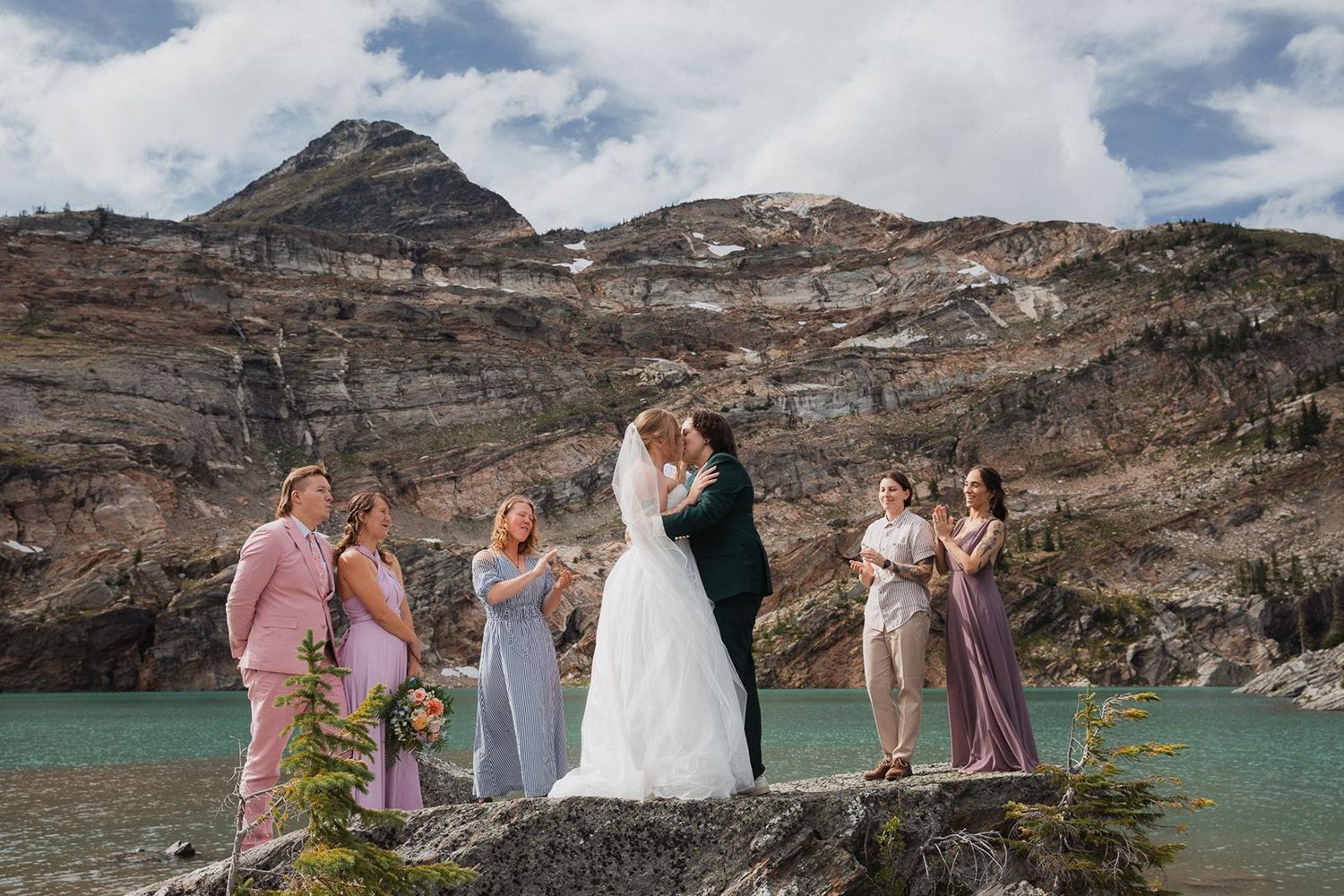 Two brides share a kiss surrounded by wedding guests beside a turquoise mountain lake.