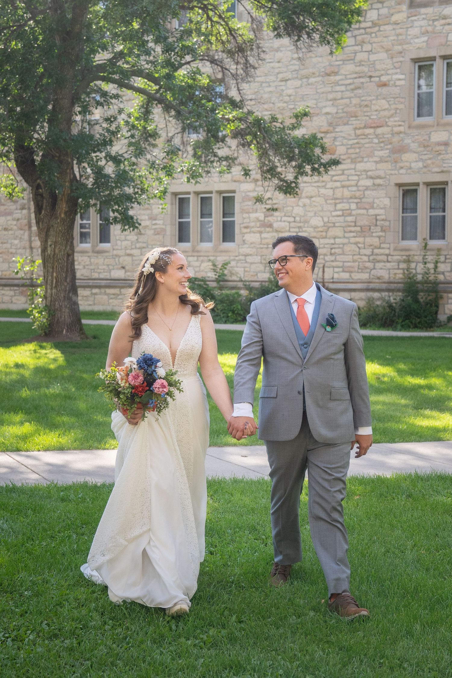 A couple in wedding attire walks and poses playfully on a grassy lawn near a stone building on their special day.