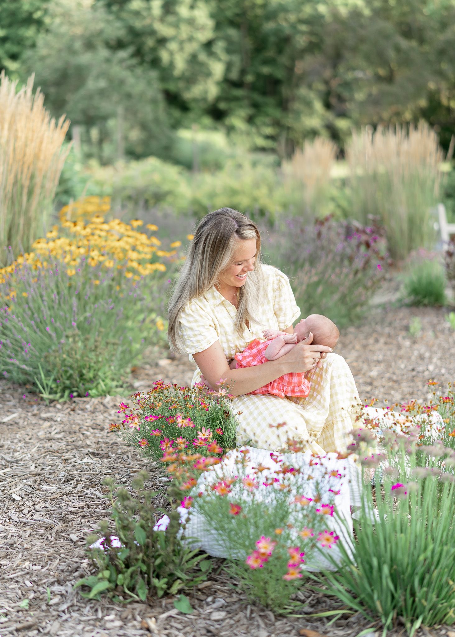 A person in a white dress sits among colorful summer flowers and ornamental grasses in a garden setting.