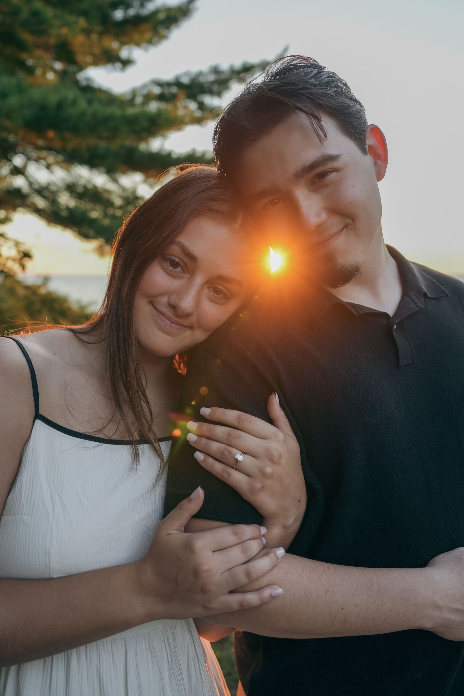 A diamond engagement ring sits on a black textured surface with champagne glasses blurred in the background.