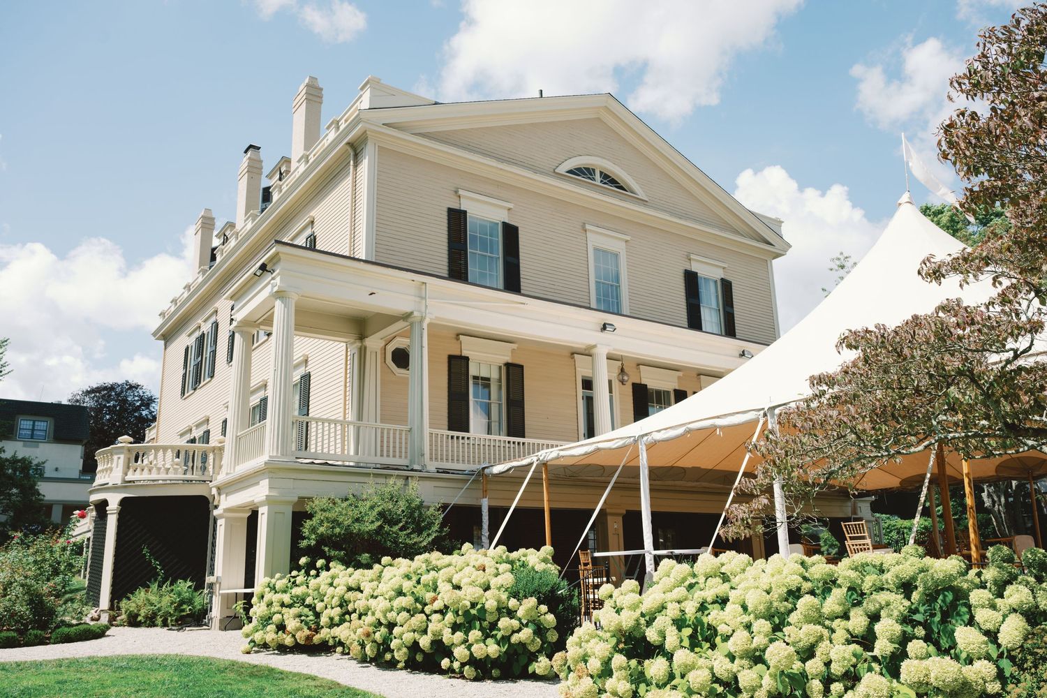 Elegant white mansion with green shutters and a sweeping tent canopy set against a bright blue summer sky.