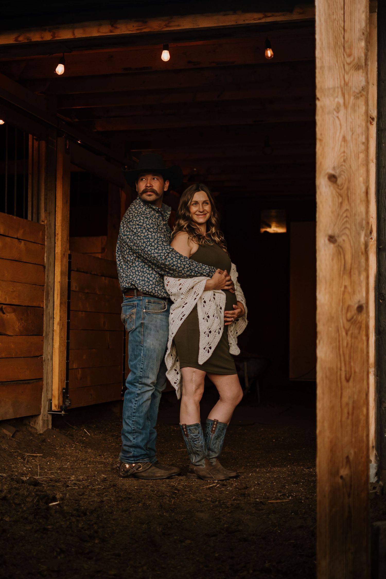A romantic couple stands together in a rustic barn setting with string lights overhead and wooden walls in the background.