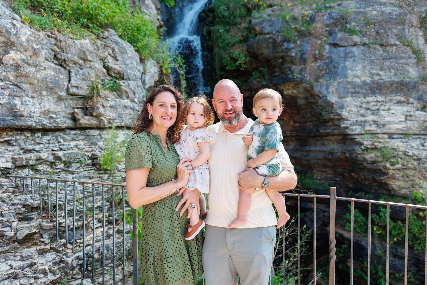 A family poses together at a scenic waterfall overlook with stone walls and metal railings in the background.