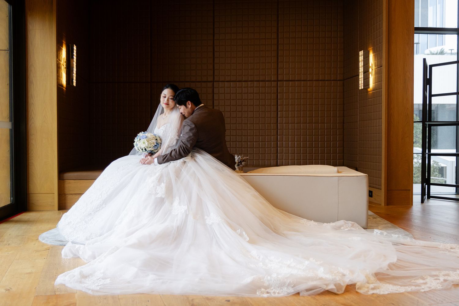 A couple embraces outdoors in wedding attire, with the bride in white lace and groom in navy suit on a sunny garden day.