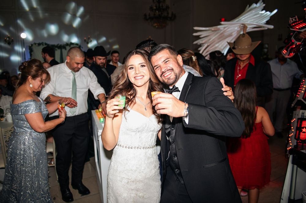 Couple celebrating at a wedding reception holding drinks under decorative lighting.