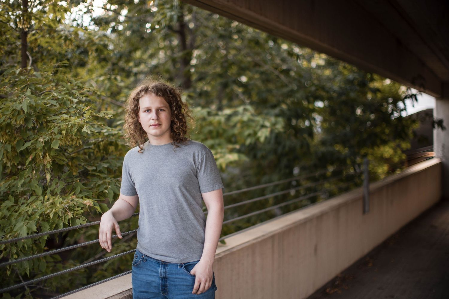 Young person with curly hair in gray t-shirt and jeans standing on an outdoor walkway surrounded by green trees.