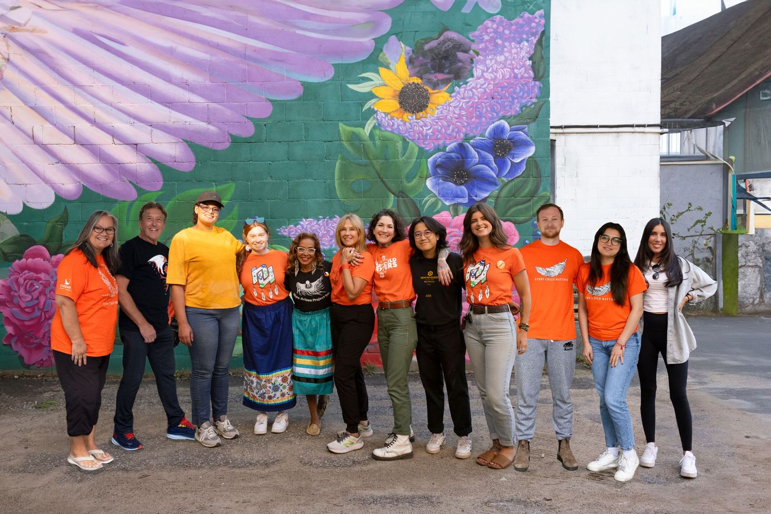Group photo of people in orange shirts standing against colorful mural with wings and sunflower design.