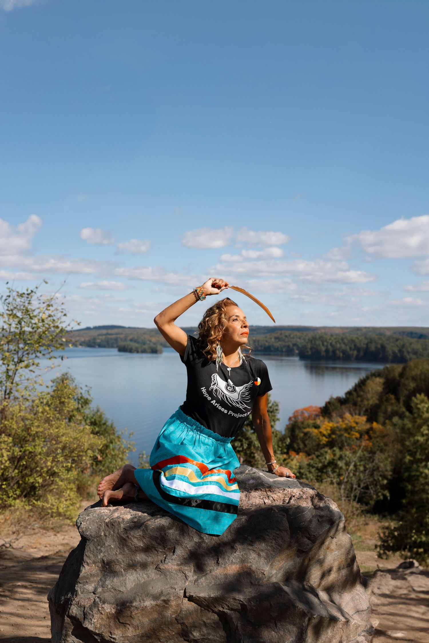 Person in turquoise dress poses dramatically on large rock formation overlooking lake.