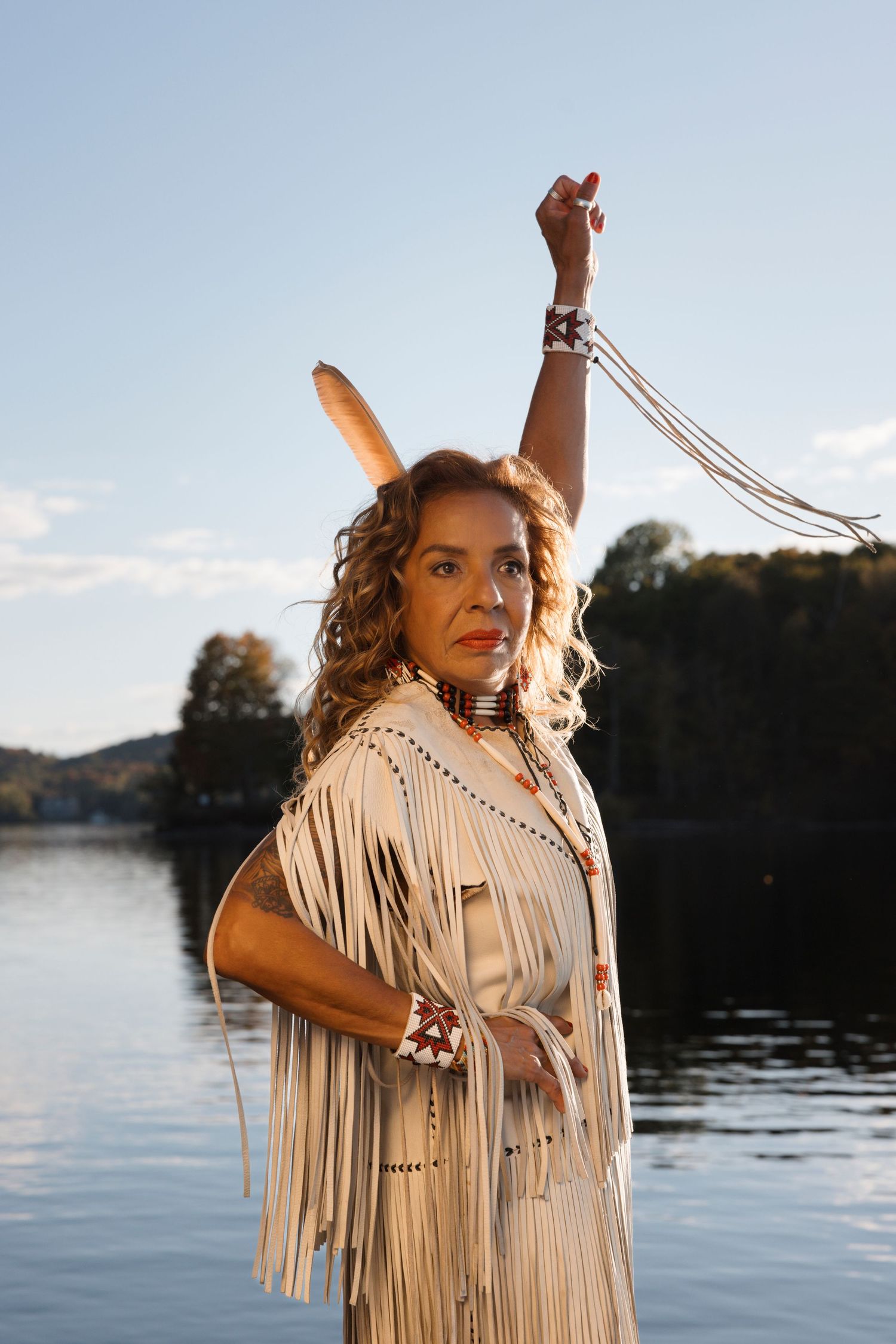 Person in fringed leather outfit poses dramatically by lakeside at sunset.
