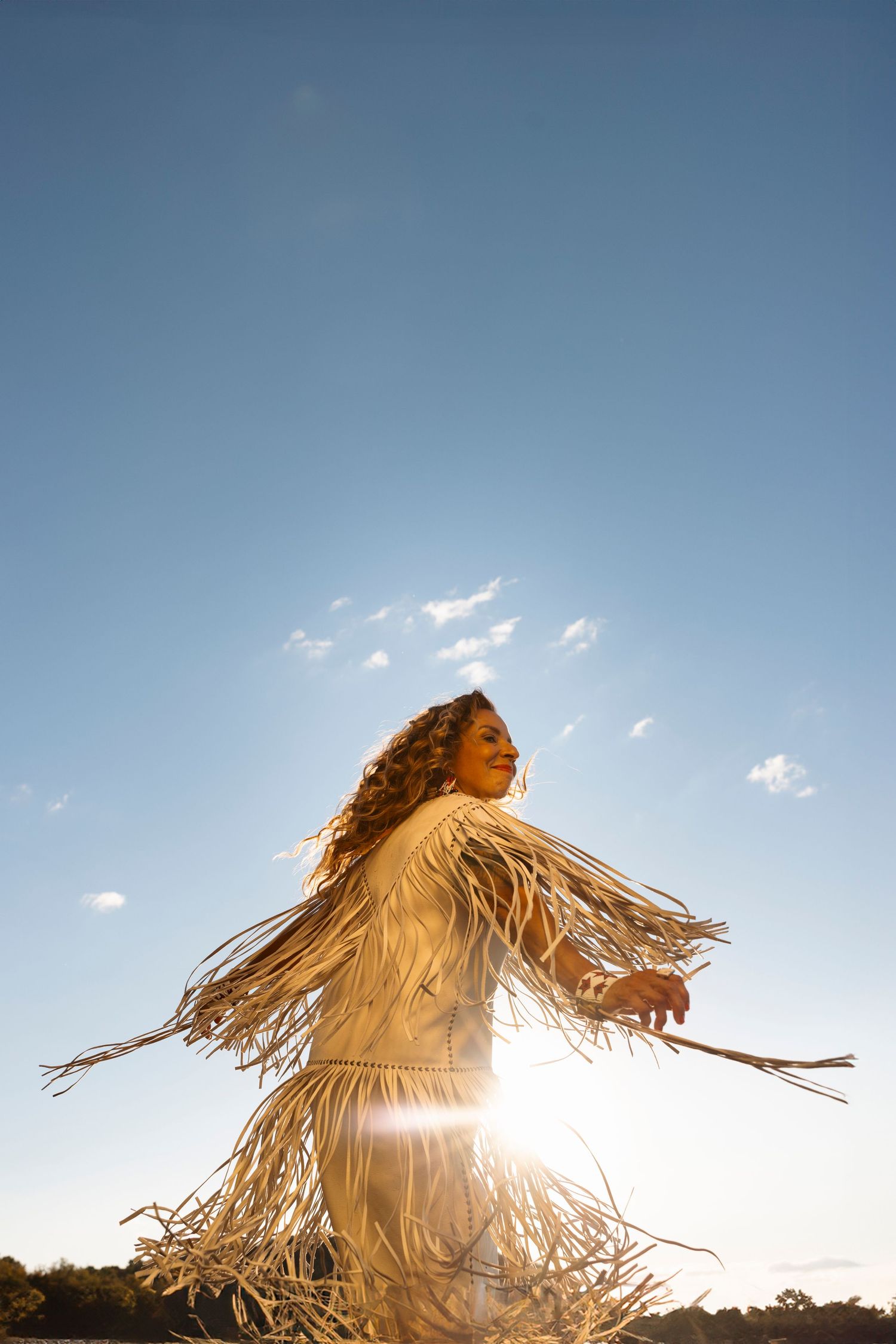 Silhouette of figure with flowing fringed garment dancing against bright sunlit sky.