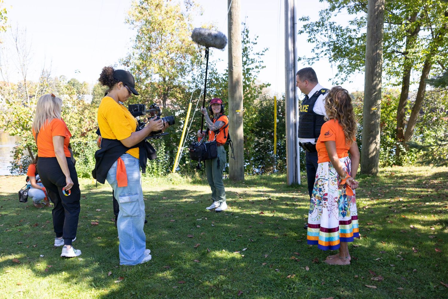 Group of people gathered outdoors in a park setting with trees and grass on a sunny day.