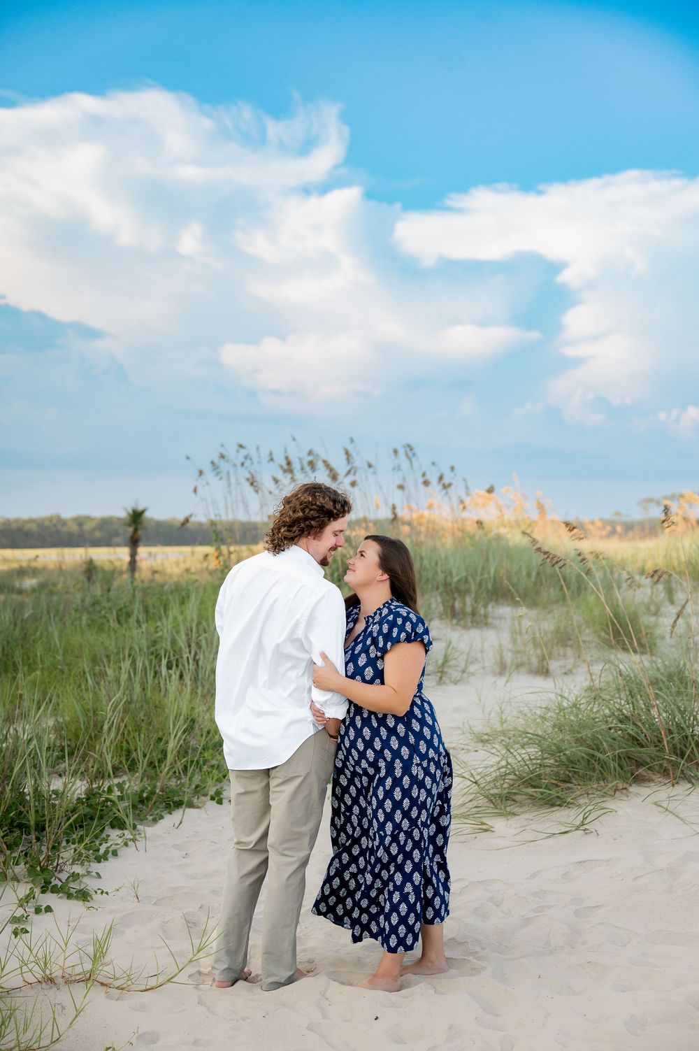 A couple shares an intimate moment on sand dunes with sea grass and blue skies in the background.