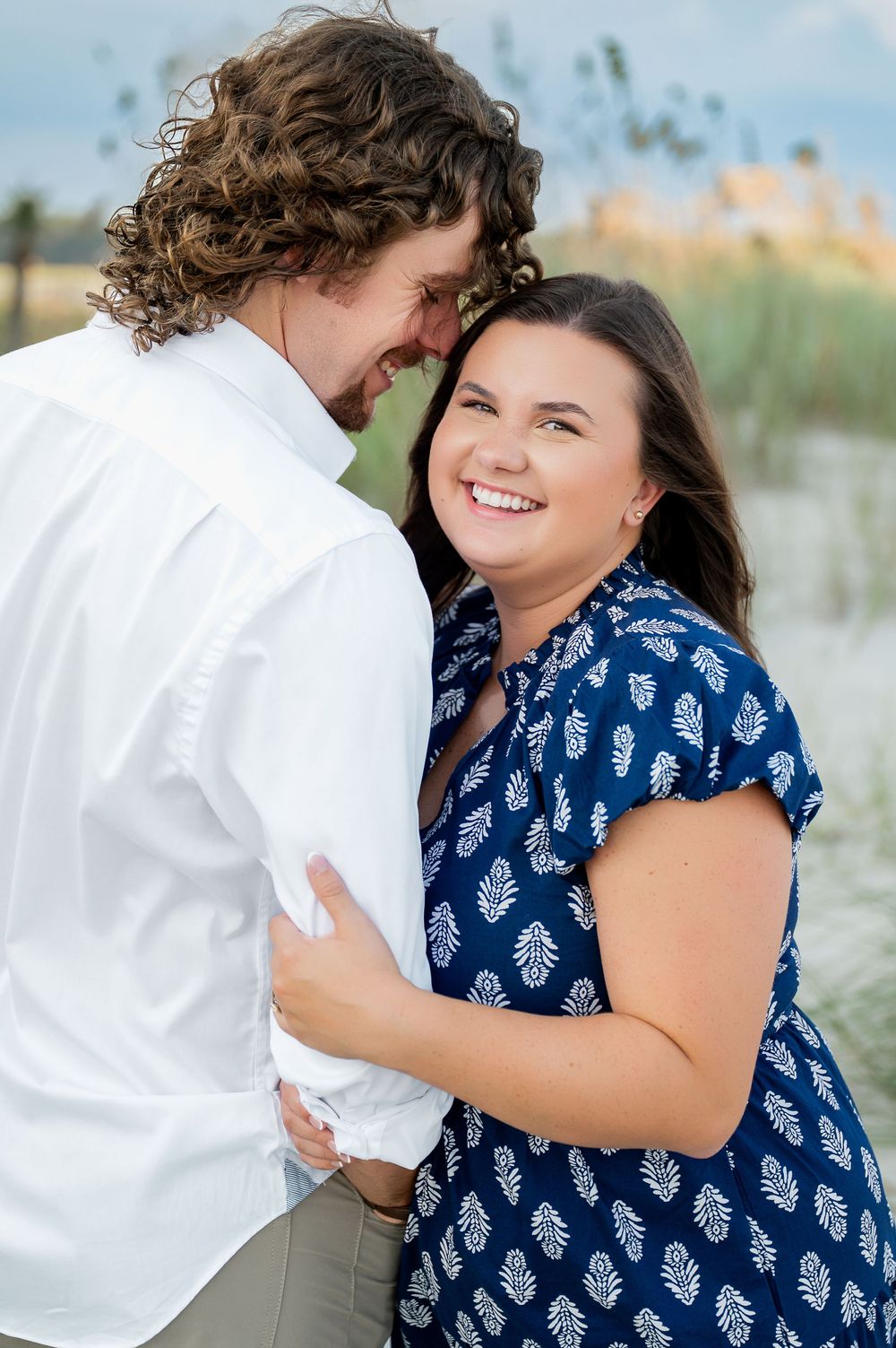 A couple in coordinated outfits shares laughs and embraces on a beach with dune grass.