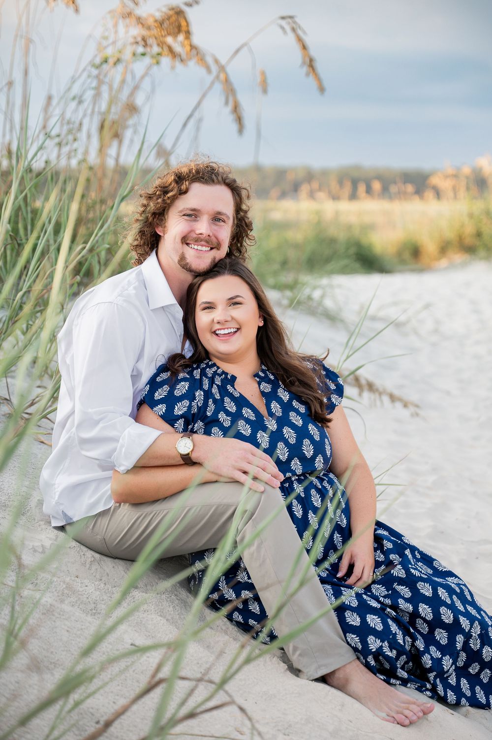 A couple relaxes together on beach dunes surrounded by coastal grass on a bright day.