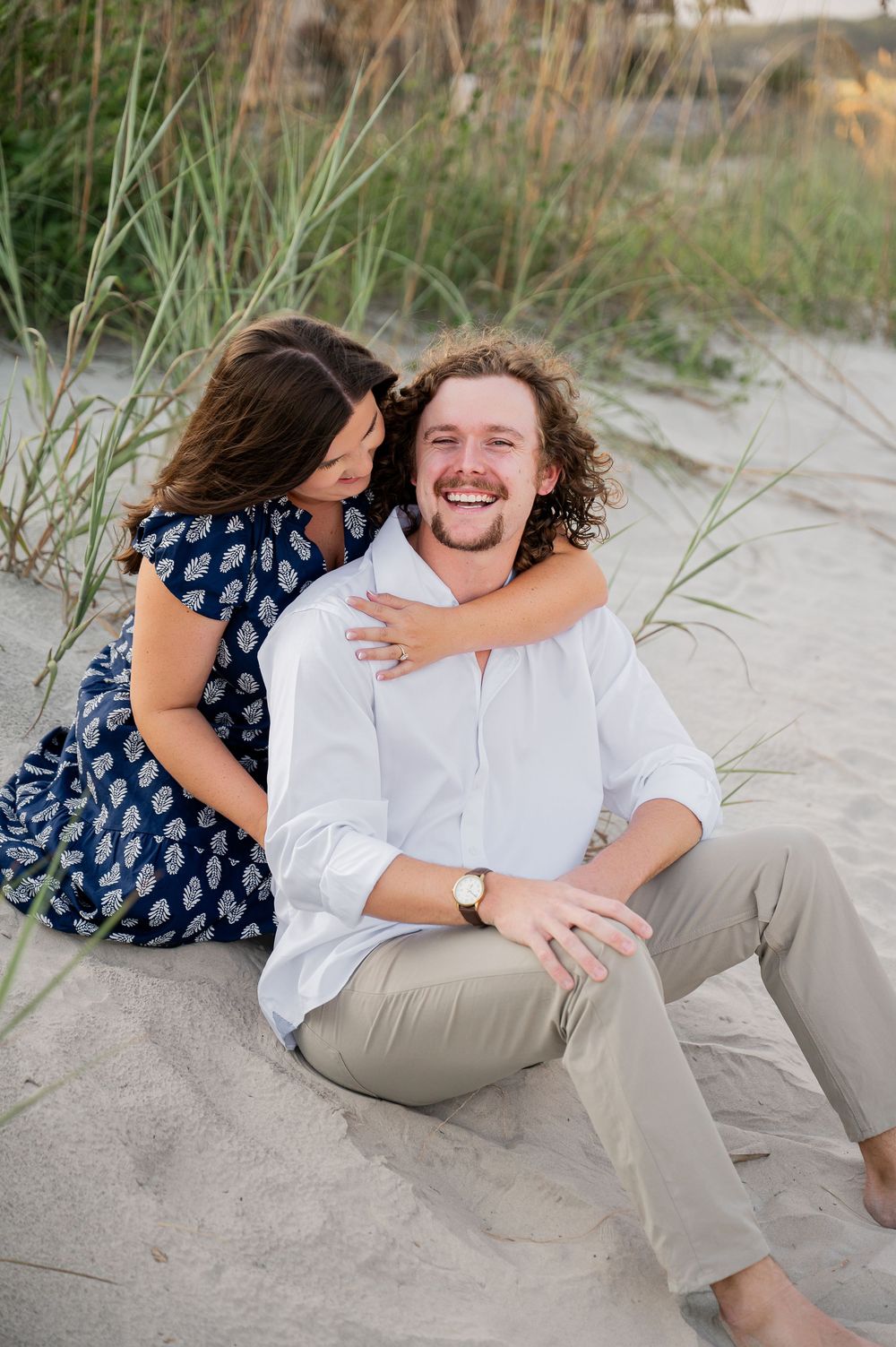 Couple laughing and embracing while lying on white sand beach during sunset.
