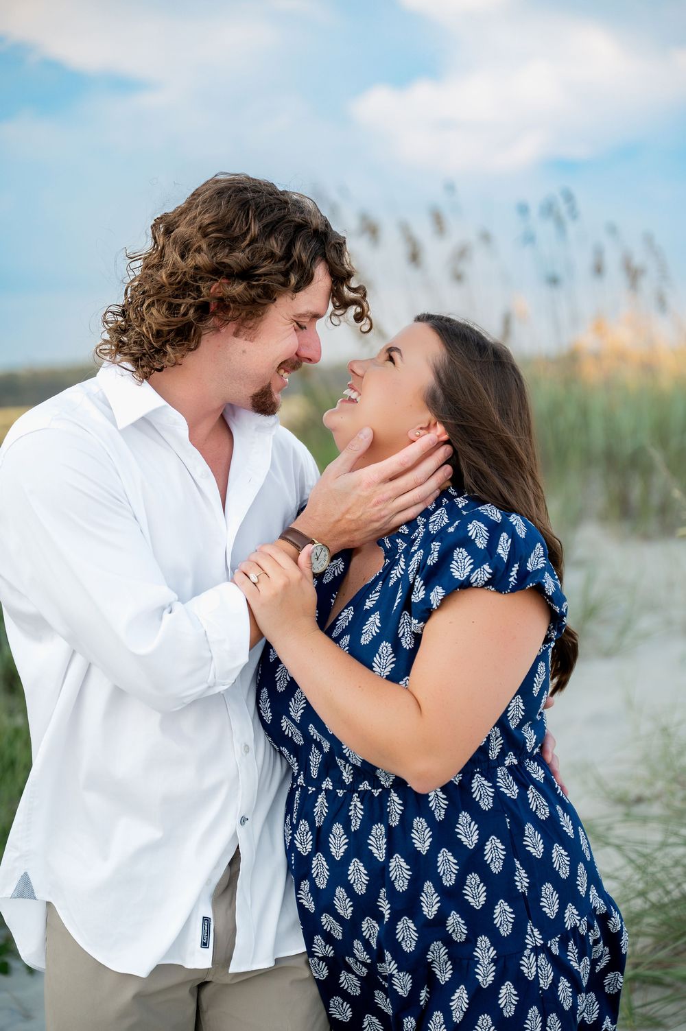 A playful couple shares intimate moments on a sandy beach with coastal vegetation in the background.