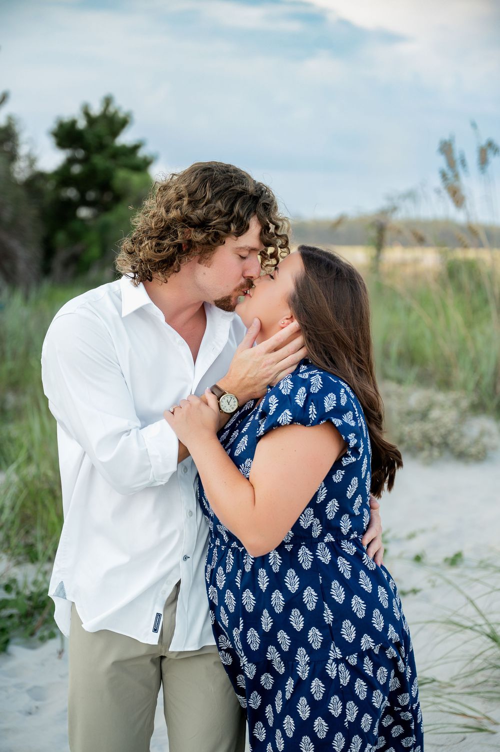 A tender moment between a couple in white and blue outfits on a sandy beach with dune grass.