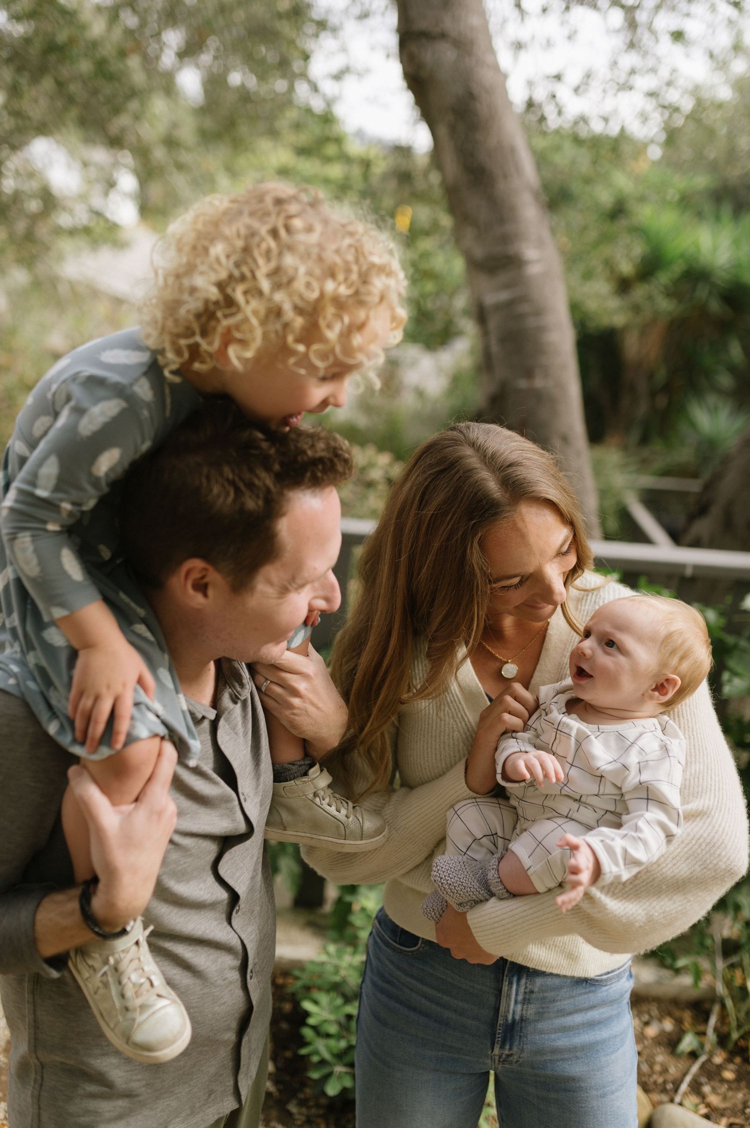 A candid family photo taken outdoors showing joyful interactions and laughter between parents and their young children.