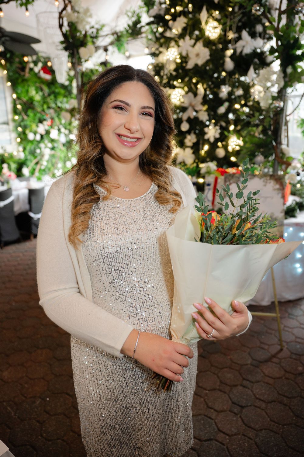 A person in a sparkly beige dress and cardigan holding a white floral bouquet against a twinkle light backdrop.