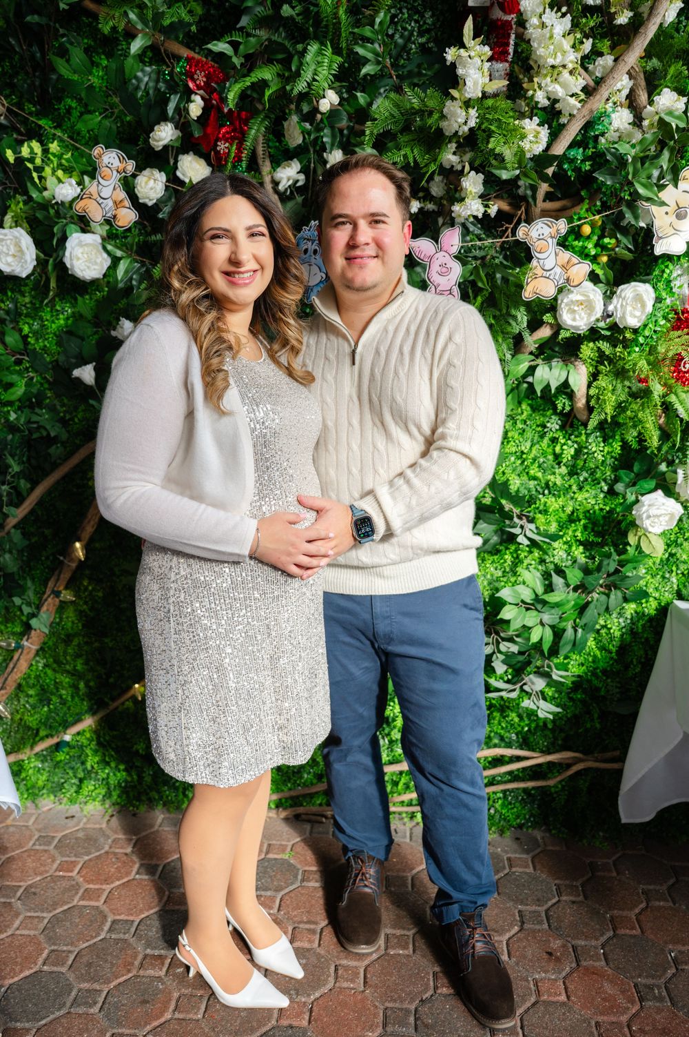 A couple in coordinating white outfits poses against a lush floral wall backdrop at an event venue.