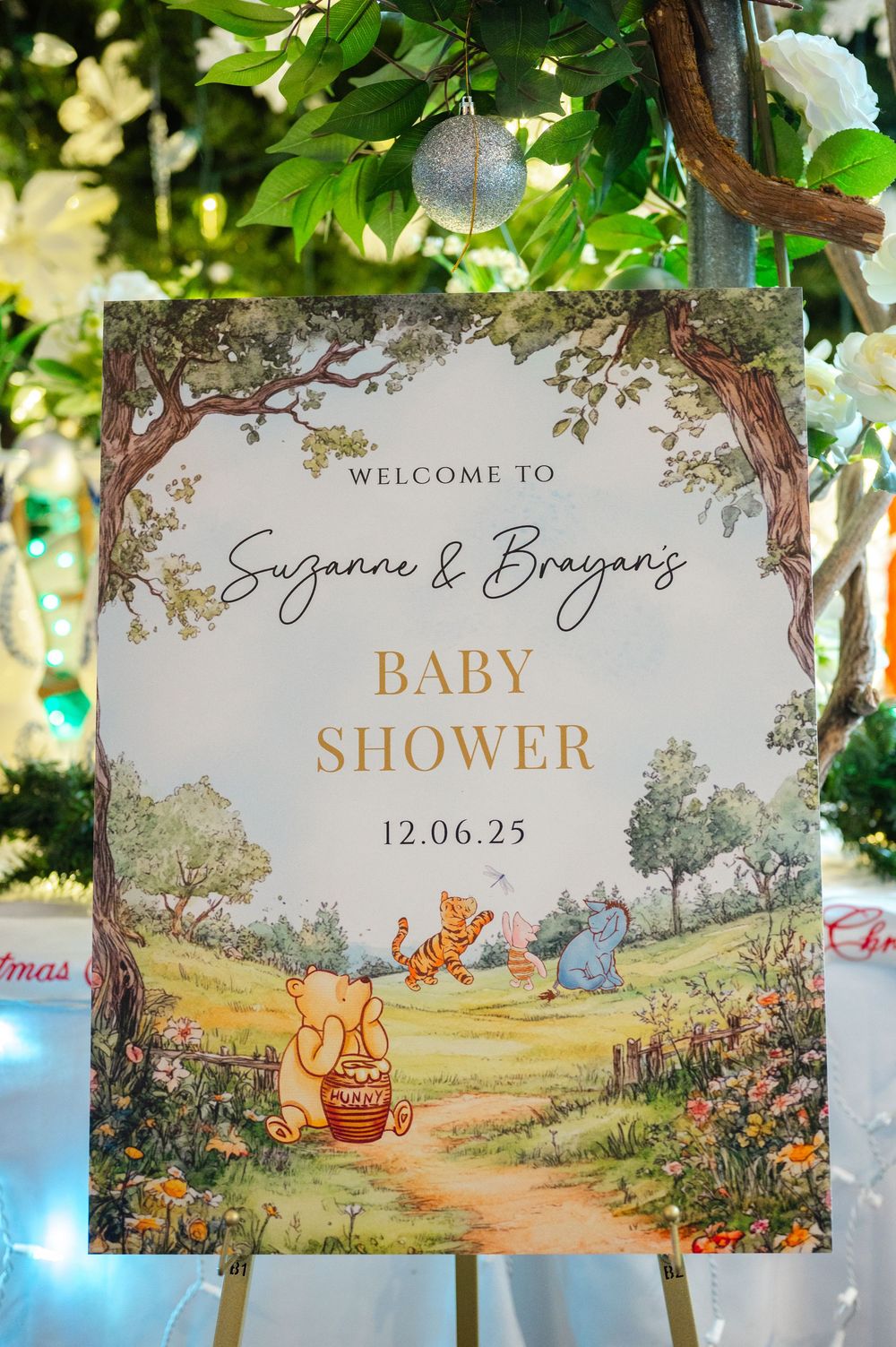 A baby shower welcome sign featuring Winnie the Pooh characters in a whimsical woodland setting with gold lettering.