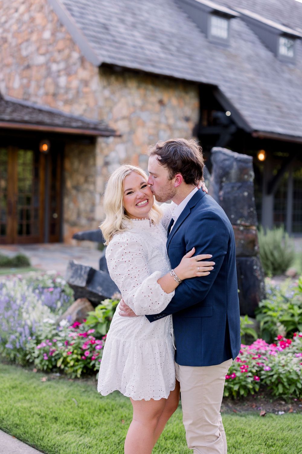 A couple shares romantic moments in front of a rustic stone building with blooming garden flowers.